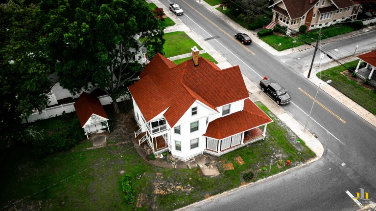 Used - Your local expert roofing company. Aerial view of a large, two-story white house with a new GAF Timberline HDZ Patriot Red Shingles roof on a corner lot in Pocomoke City, MD. It features a wraparound porch and abundant greenery. Two cars are parked nearby, with neighboring houses in sight, expertly roofed by G and Bros Roofing LLC. G and Bros Roofing LLC Roofing Contractor Located in Salisbury MD.