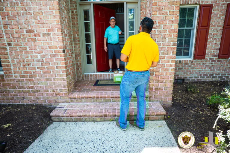 Used - Your local expert roofing company. A person in a yellow shirt delivers a package to someone at the doorway of a brick house, embodying our "Why We Don't Want to Replace Your Roof: G and Bros Roofing Perspective." The recipient stands near a green box, with red shutters and a welcome mat. Two logos are visible in the bottom right corner. G and Bros Roofing LLC Roofing Contractor Located in Salisbury MD.