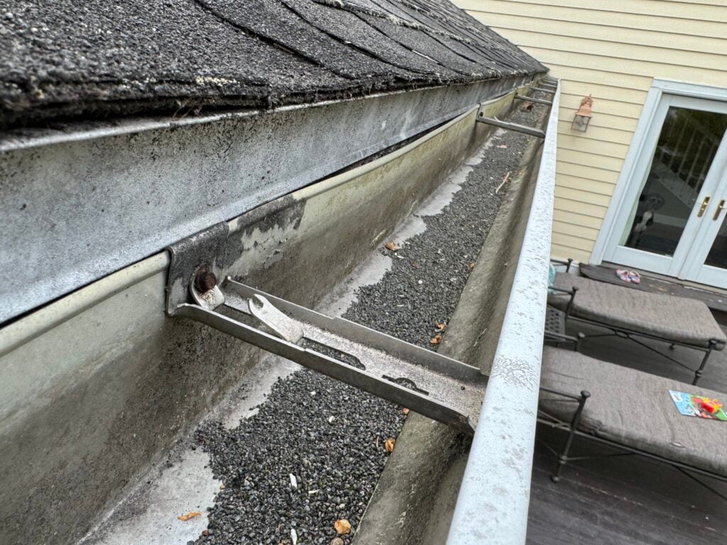 Used - A close-up shot of dark shingle granules pooled at the end of a white gutter downspout or on a splash block. Salisbury MD G and Bros Roofing