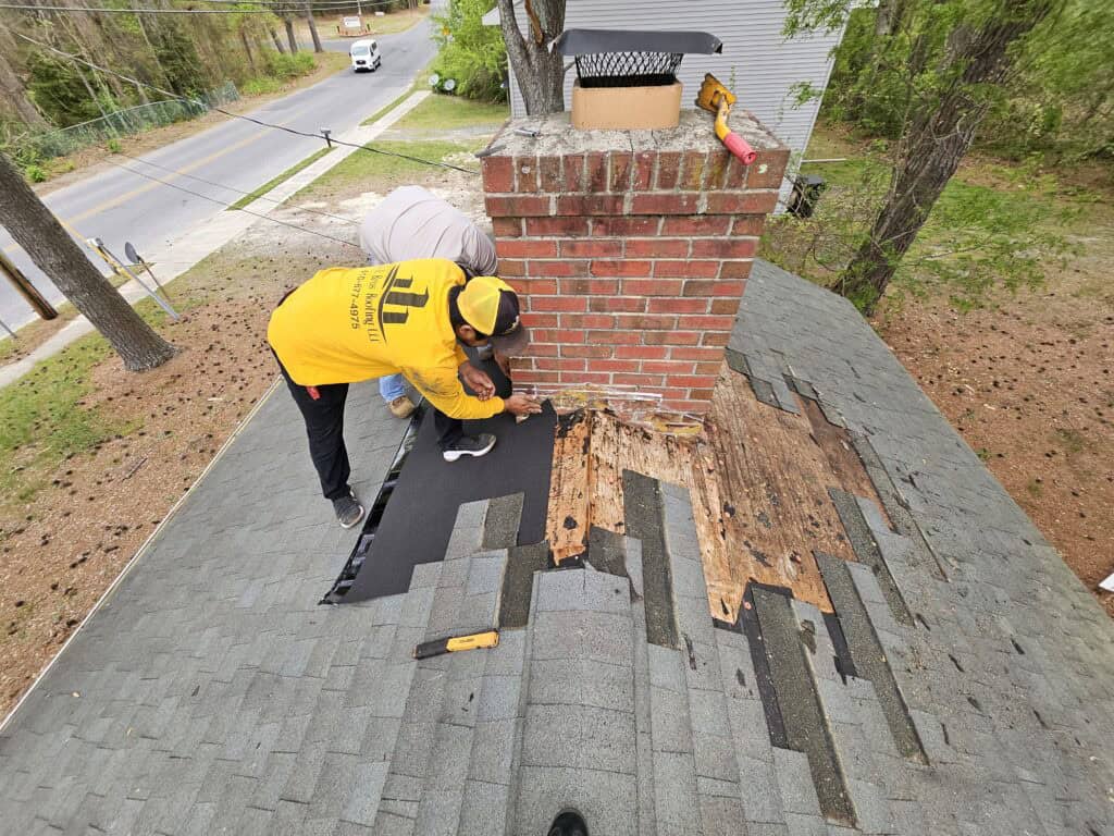 Used - G and Bros employees installing new chimney flashing on a residential asphalt shingle roof with exposed roof decking around a brick chimney