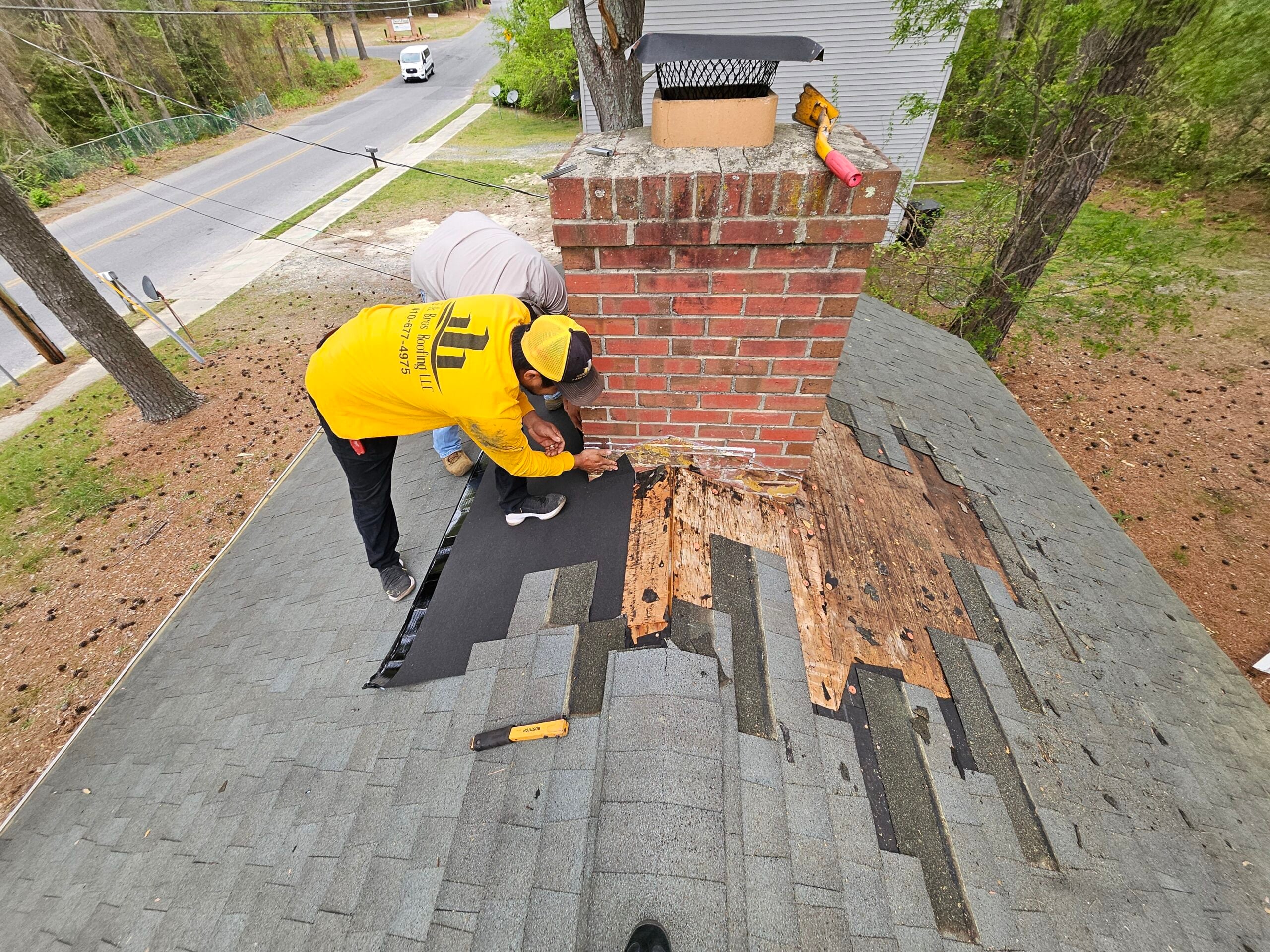G and Bros technician on a residential roof inspecting shingles near a chimney flashing. Bright morning light, close-up real job photo, technician in company shirt or vest.